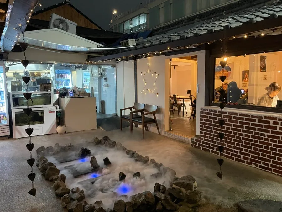 밤의 수상한한옥 마당 — 기와 처마의 전구 조명과 안개 낀 돌 연못, 안쪽 실내 좌석